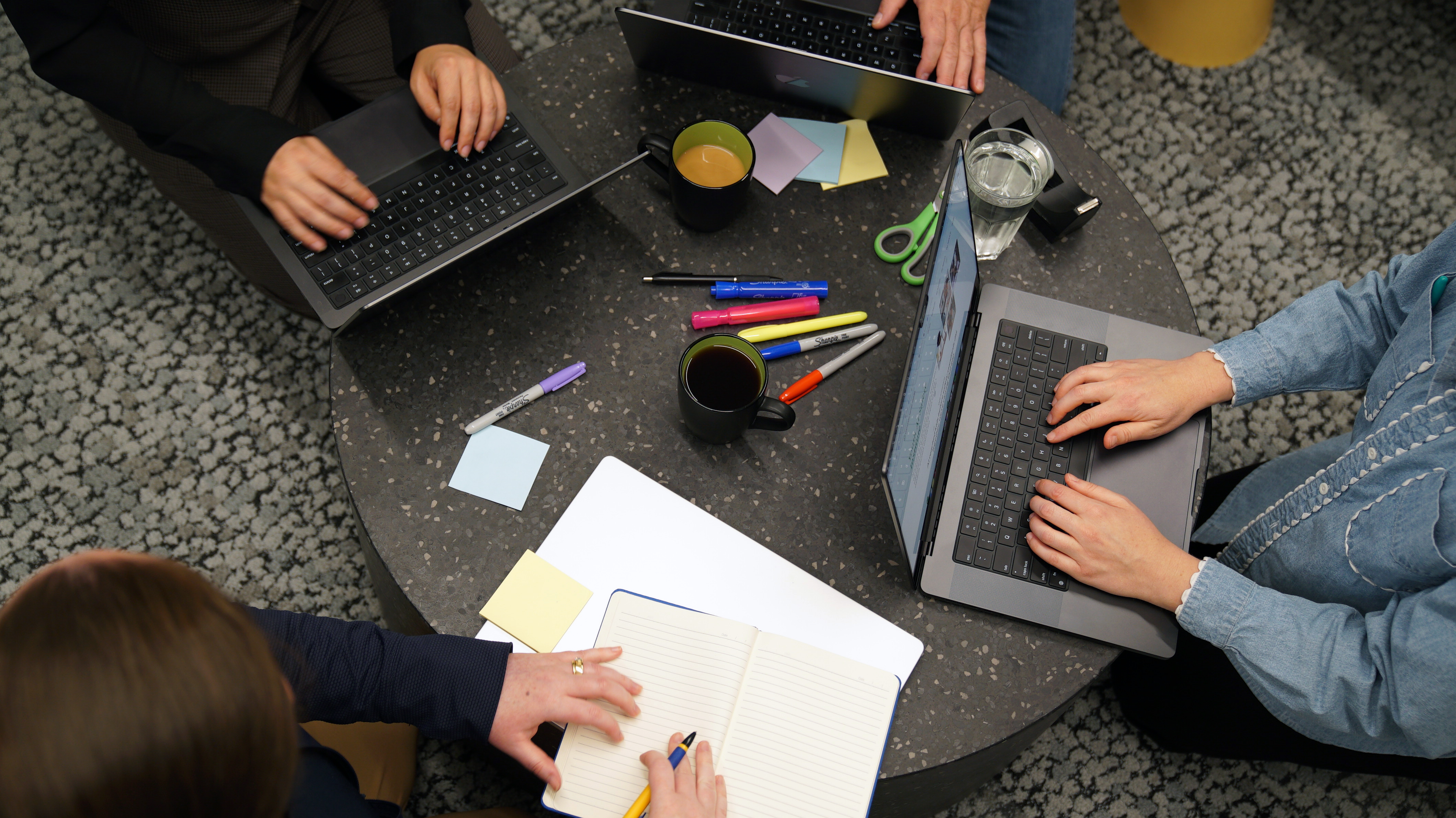 A top-down photo of a table with four individual working, three using laptops and one using a notebook